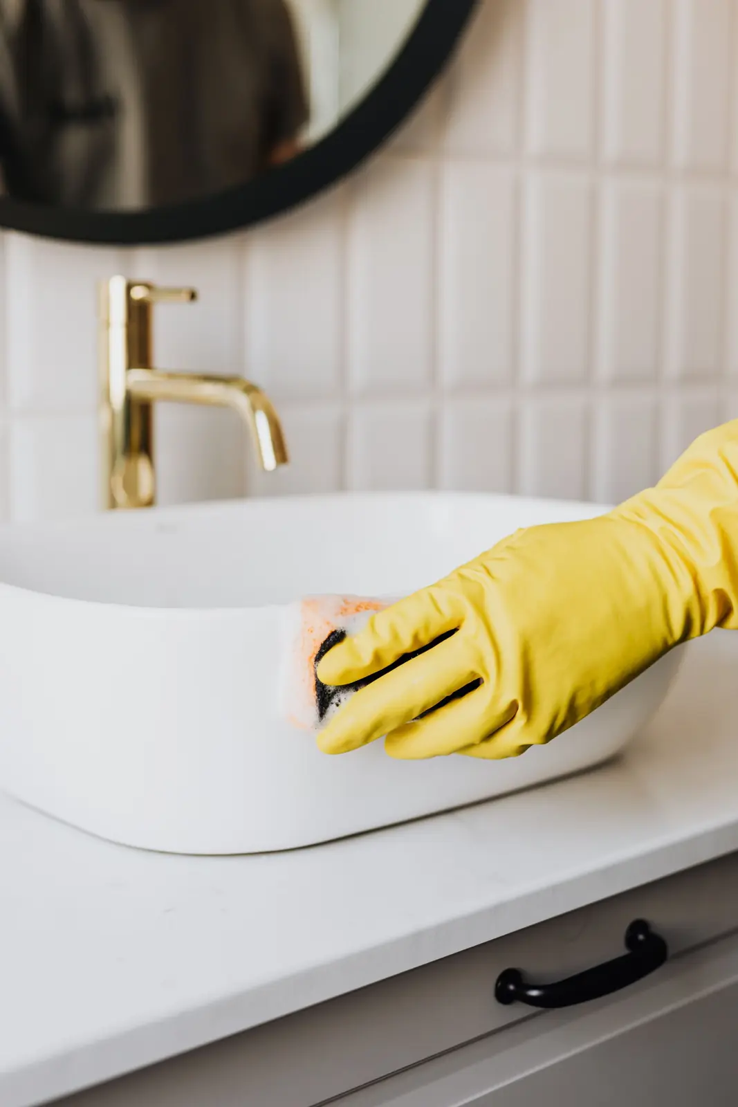 A gloved hand cleaning a white bathroom sink with a brass faucet